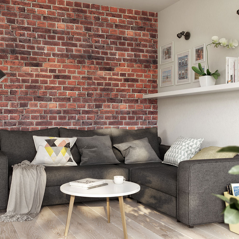 Modern living room with a dark gray couch, decorative pillows, exposed brick wall, floating shelf with frames, round coffee table with a book and coffee cup, and sunlight casting shadows on the wooden floor.