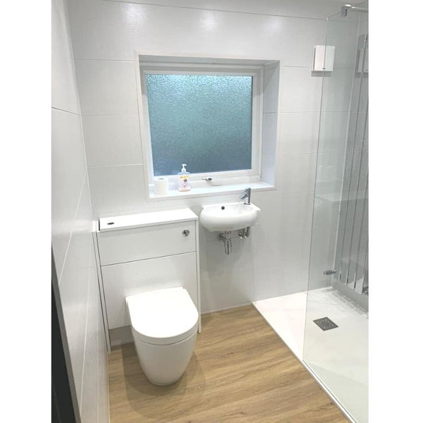 Modern bathroom interior with white tiles, frosted glass window, wall-mounted sink, white toilet, wooden floor, and clear glass shower enclosure.