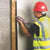 Construction worker applying pink adhesive from a Pinkgrip cartridge to secure wooden frame on concrete wall, wearing safety gear including hard hat and reflective vest
