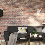 Modern living room with brick wall, dark gray sofa with decorative pillows, round coffee table with plants, cozy blanket, geometric wall art, and a white deer head wall decor.