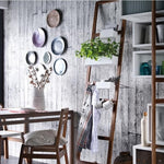 Modern rustic dining area with wooden furniture, decorative ceramic plate wall display, ladder shelf with green plants, kitchen accessories, and distressed white wooden wall background.