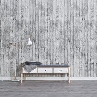 Modern minimalist interior with textured grey wooden plank wallpaper, sleek white and wood bench with storage drawers, cozy gray cushion and throw blanket, stylish floor lamp with white shade and wooden accents, and gray flooring.