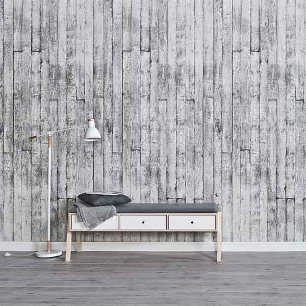 Modern minimalist interior with textured grey wooden plank wallpaper, sleek white and wood bench with storage drawers, cozy gray cushion and throw blanket, stylish floor lamp with white shade and wooden accents, and gray flooring.