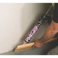 Construction worker applying adhesive with PINKGRIP caulk gun to secure wooden baseboard on a wall during interior finishing work
