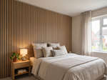 A modern bedroom featuring a bed dressed in neutral beige linens, set against a prominent wall with vertical wooden slats. A light wood nightstand with a lit table lamp sits beside the bed, and a window with sheer curtains is visible on the right.