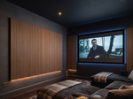 A home theater room with a large screen displaying a movie, set in a dark room with wood slat accent walls, a dark sectional sofa, and a coffee table in the foreground.