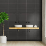 Modern bathroom interior with double sink, matte black faucets, dark wall tiles, wooden shelf, decorative plant, and stacked toilet paper rolls.