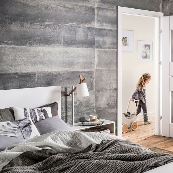 Child playing with toy stroller in a modern bedroom with a gray textured wall, white bed linens, wooden floor, and open door.