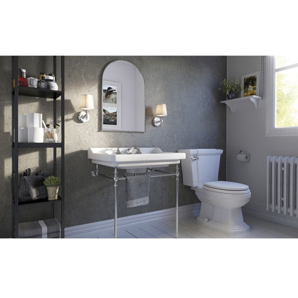 Modern bathroom with textured gray walls, white pedestal sink and toilet, black shelving unit with towels and toiletries, wall-mounted mirror, sconce lighting, and houseplants.