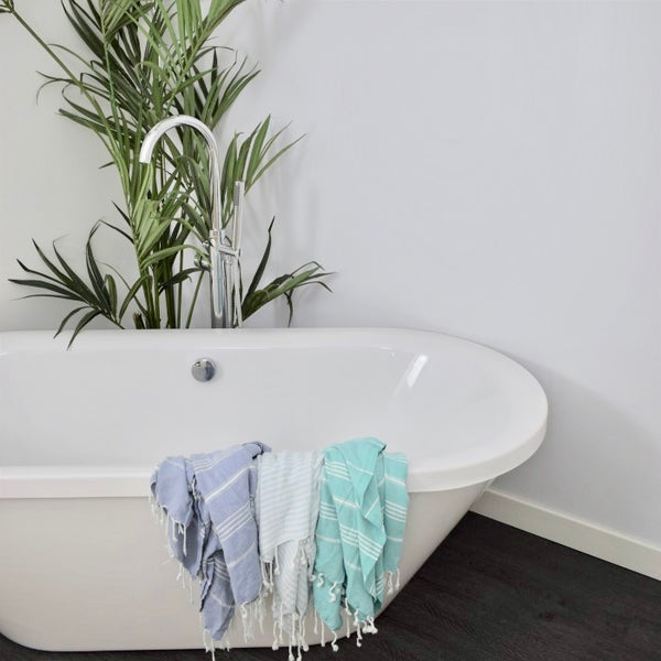 Modern bathroom with freestanding white bathtub, elegant faucet, green potted plant and two hanging striped towels in blue and turquoise colors, on a minimalist grey floor.