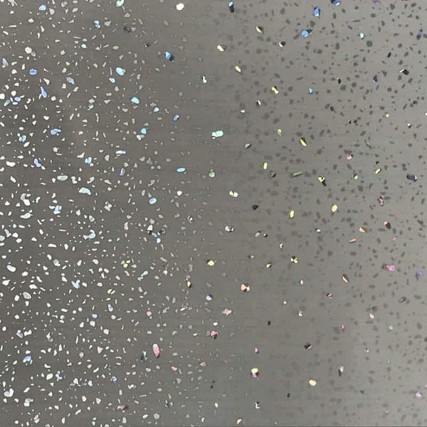Close-up of a gray surface speckled with colorful glitter, featuring a variety of sparkly specks and holographic confetti scattered across a neutral background.
