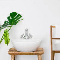 Modern bathroom interior with white vessel sink on wooden stand, silver faucet, monstera plant, and fluffy towel on towel rack against a clean white wall.