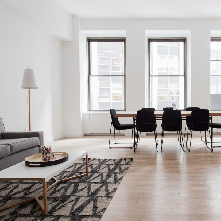 Modern minimalist living room with large windows, hardwood floors, gray sofa, white coffee table, black dining chairs, wooden dining table, patterned area rug, and floor lamp.