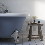 Modern bathroom interior with freestanding bathtub, chrome claw-foot detail, rustic wooden stool, decorative plant, soap dispenser, and grey wall background.