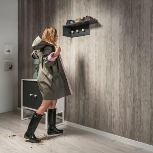 Woman in raincoat and boots placing keys on wall-mounted shelf with coat hooks in modern interior entryway