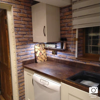 Rustic kitchen interior with brick backsplash, white cabinets, wooden countertop, and vintage dishwasher, including kitchen accessories and a patterned oven mitt on the counter.