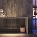 Modern bathroom interior with dark gray textured walls, concrete countertop, folded towels, decorative bottle, and woven basket under atmospheric lighting near glass door.