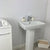 Modern bathroom interior with white ceramic pedestal sink, faucet, soap dispenser, wicker basket, folded towels on vanity, and marble tile flooring.